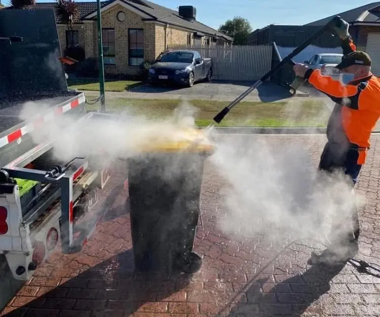 The Bin Doctor blasting a wheelie bin clean with a high-pressure hot-water wand, steam rising